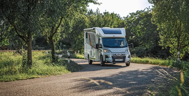 Wohnmobil der Marke Bürstner fährt auf einer ländlichen Straße durch eine grüne Landschaft.