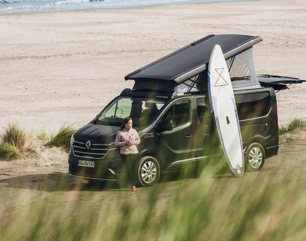 Ein Campervan Bürstner Playa mit Surfbrett steht am Strand, daneben eine Person.