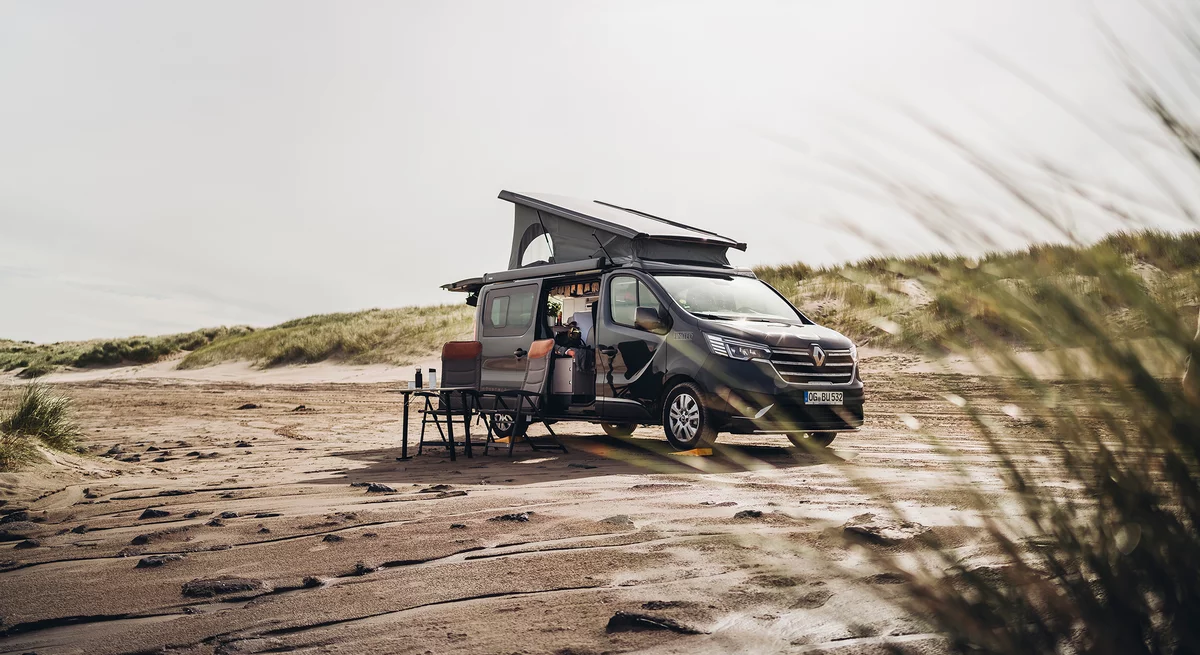 Ein Renault-Campervan steht mit geöffnetem Dach und Campingausrüstung an einem sandigen Strand.