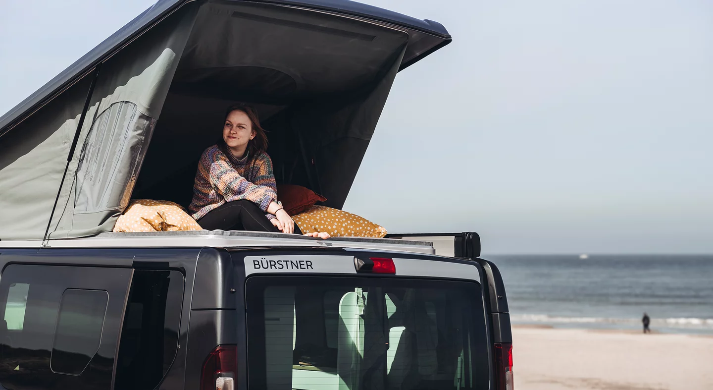 Eine Frau sitzt lächelnd im geöffneten Dachzelt eines Bürstner-Campers am Strand.