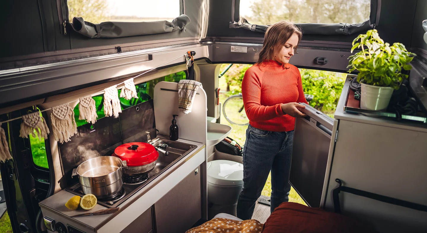 Eine Frau in einem roten Pullover steht in der Küche eines Wohnmobils und öffnet den Kühlschrank.