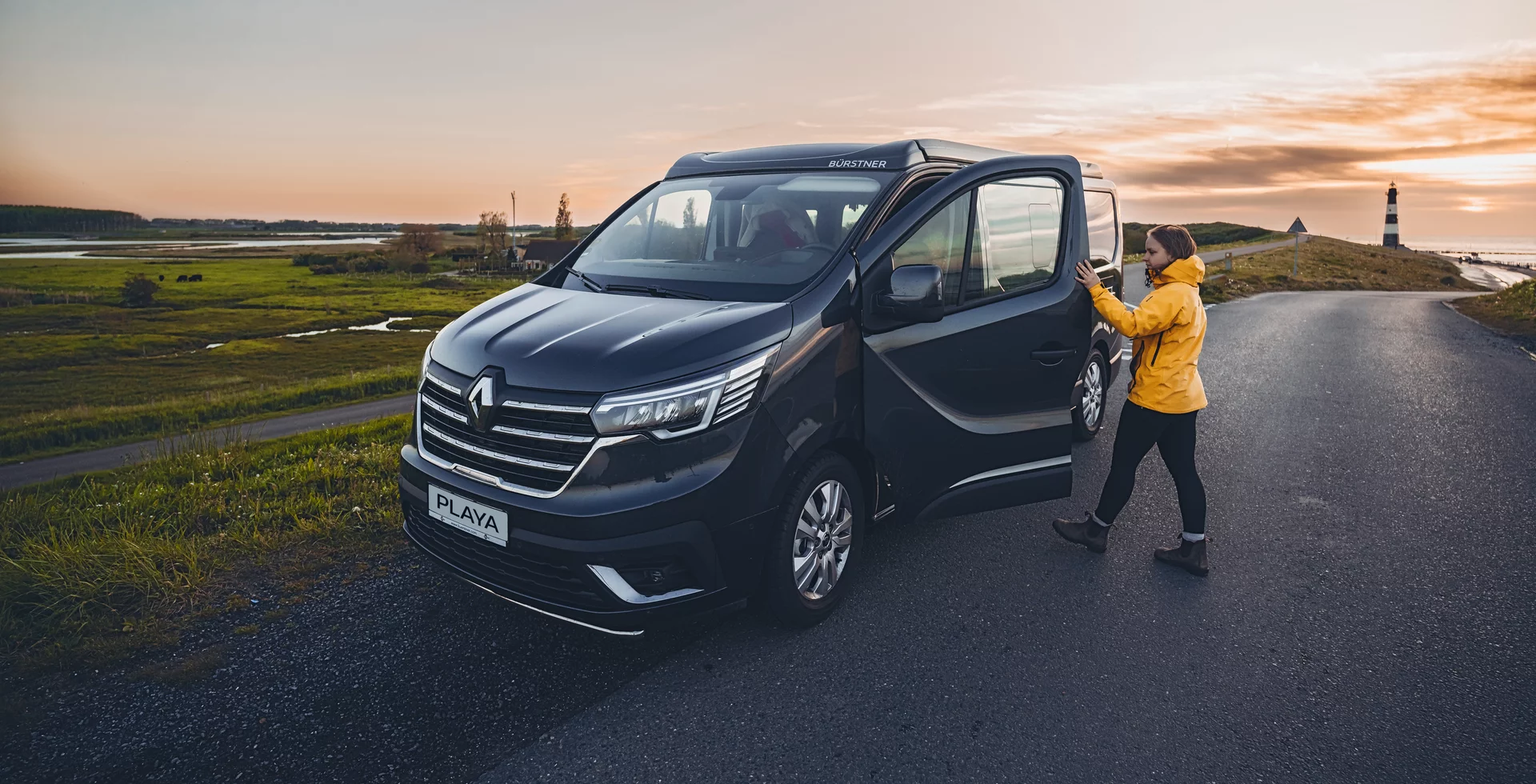 Eine Person in gelber Jacke öffnet die Tür eines schwarzen Bürstner Camper mit der Aufschrift "Playa" in einer ländlichen Umgebung bei Sonnenuntergang.