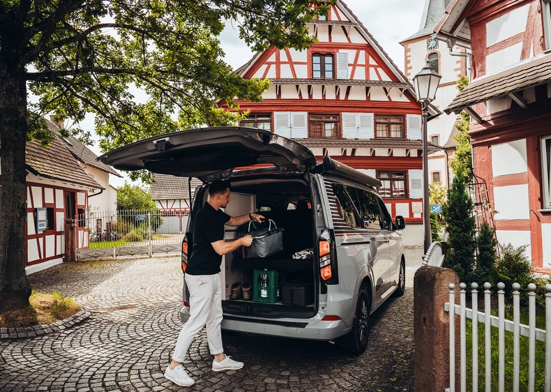 Ein Mann lädt Gegenstände in einen Ford Campervan vor einem Fachwerkhaus ein.