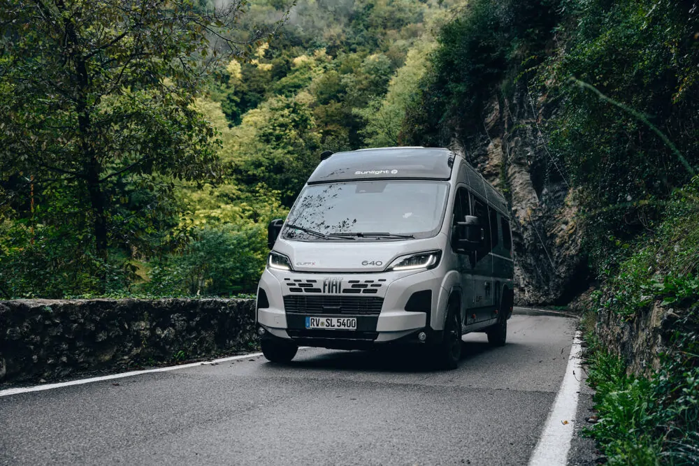 Ein Sunlight CLIFF X Wohnmobil fährt auf einer kurvigen Straße durch eine bewaldete Landschaft.