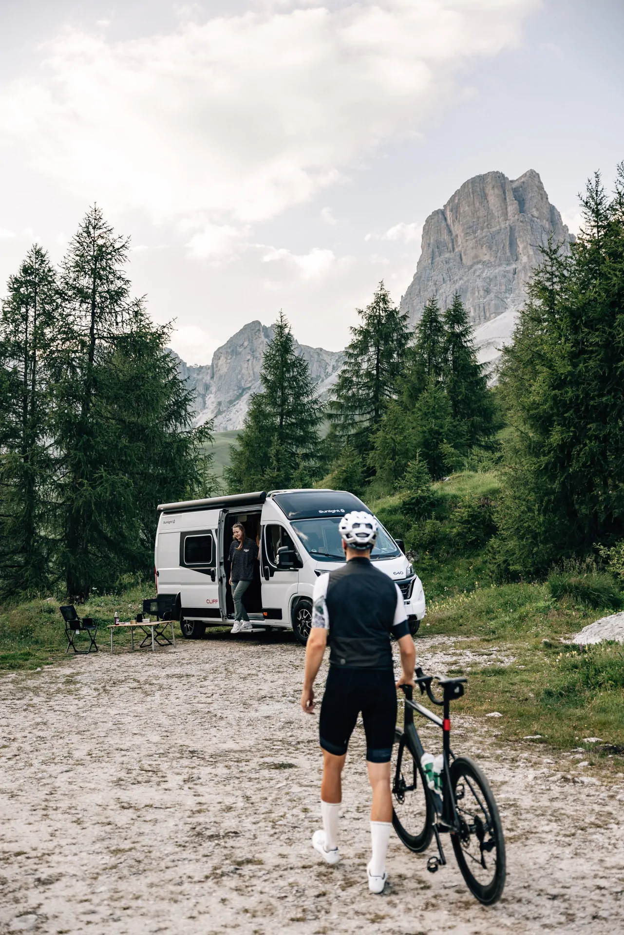 Ein Fahrradfahrer steht vor einem geparkten Sunlight CLIFF X Camper in einer bergigen Waldlandschaft.