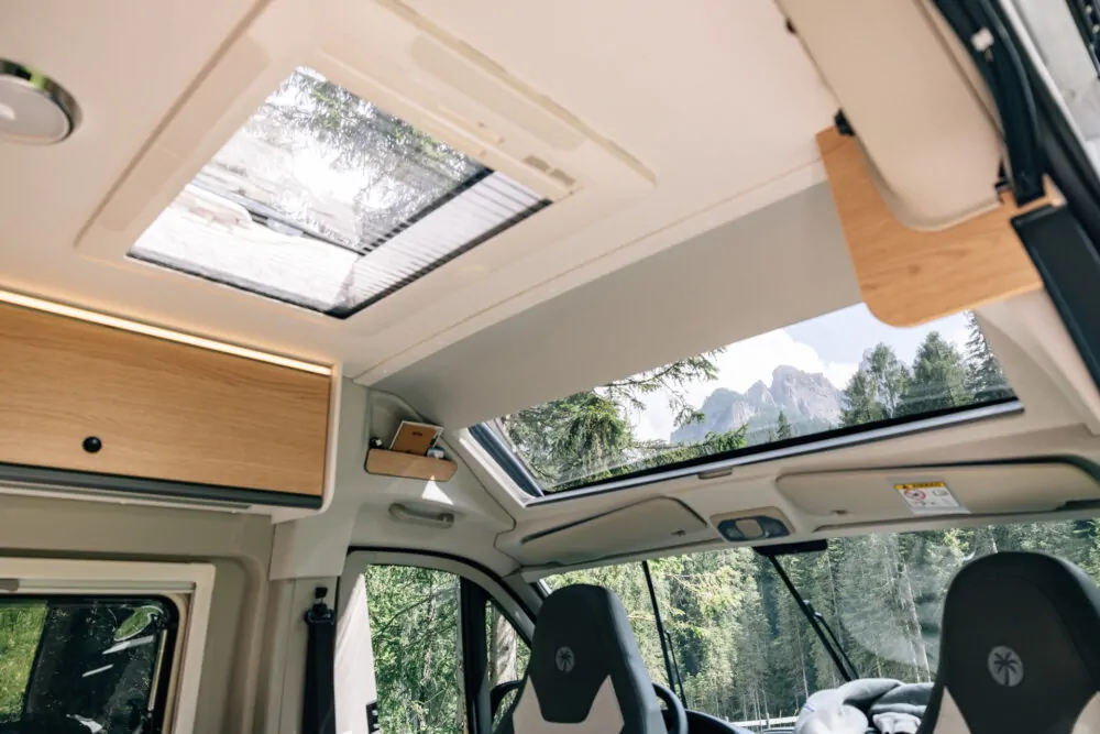 Innenansicht eines Campervans mit großem Dachfenster und Blick auf bewaldete Berge.
