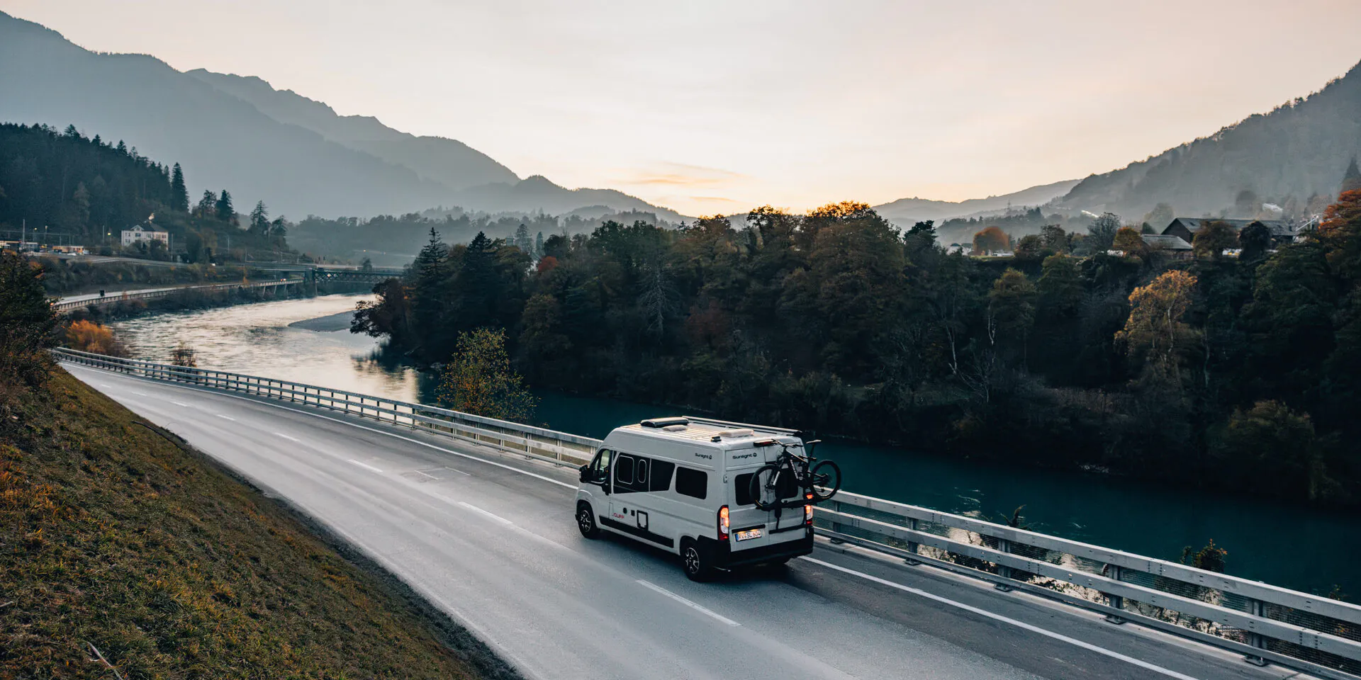 Ein weißer Campervan Sunlight Cliff fährt auf einer Straße entlang eines Flusses in einer bergigen Landschaft bei Sonnenuntergang.