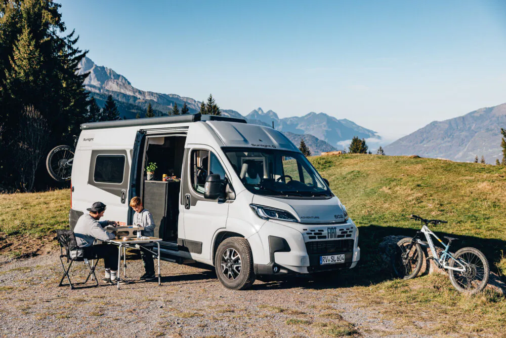 Zwei Personen sitzen an einem Tisch vor einem Fiat Sunlight Cliff Adventure Wohnmobil in bergiger Landschaft.