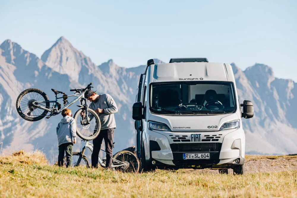 Zwei Personen laden Fahrräder auf einen FIAT Sunlight Cliff Adventure 602 Camper vor Bergkulisse.