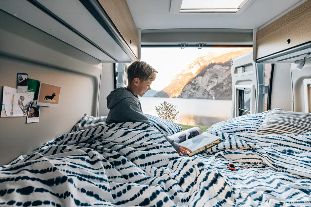 Eine Person liest in einem Bett im Inneren eines Wohnmobils mit Blick auf einen See und Berge.