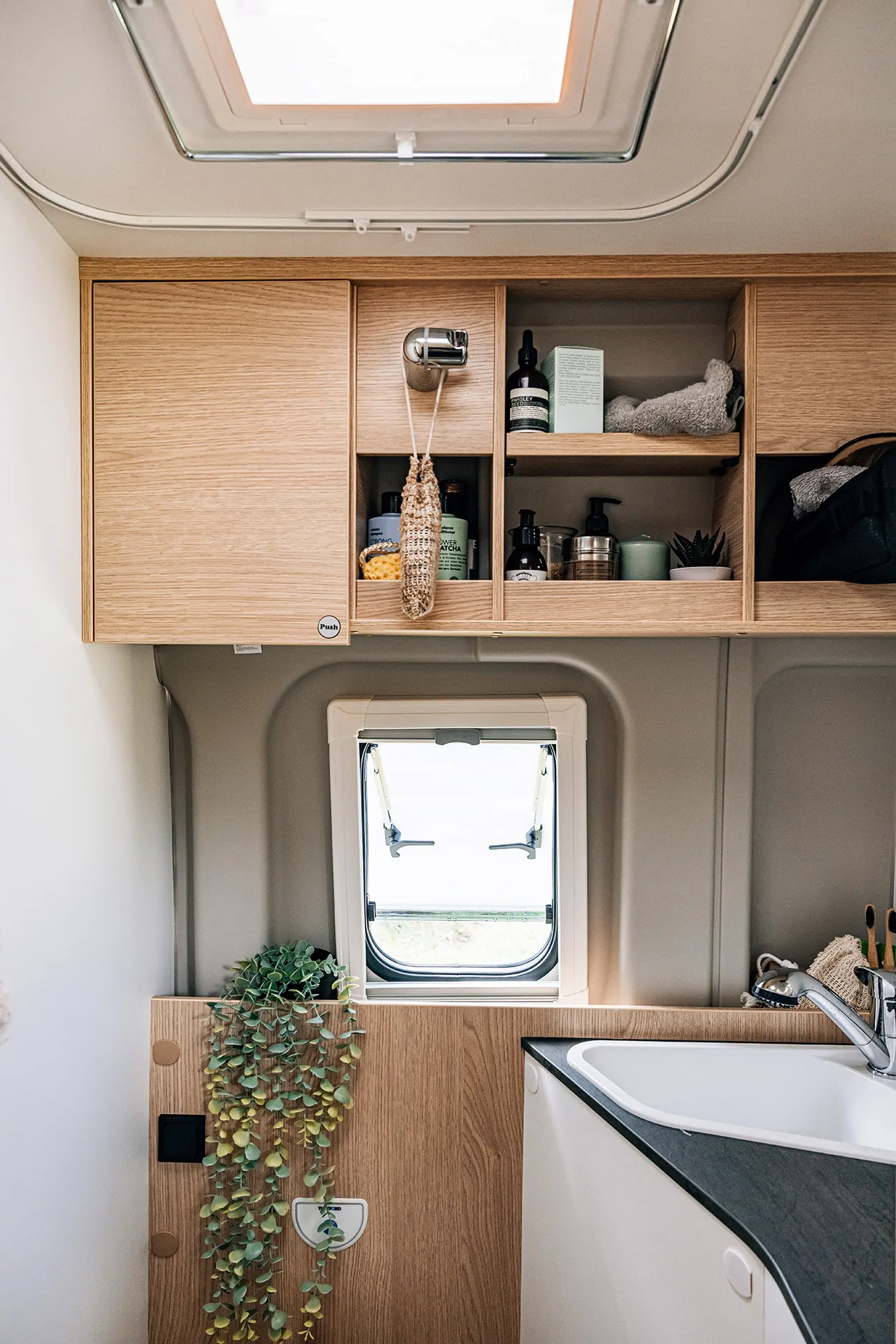 Innenansicht eines Camper-Bads mit Holzregalen, Waschbecken und kleinem Fenster.