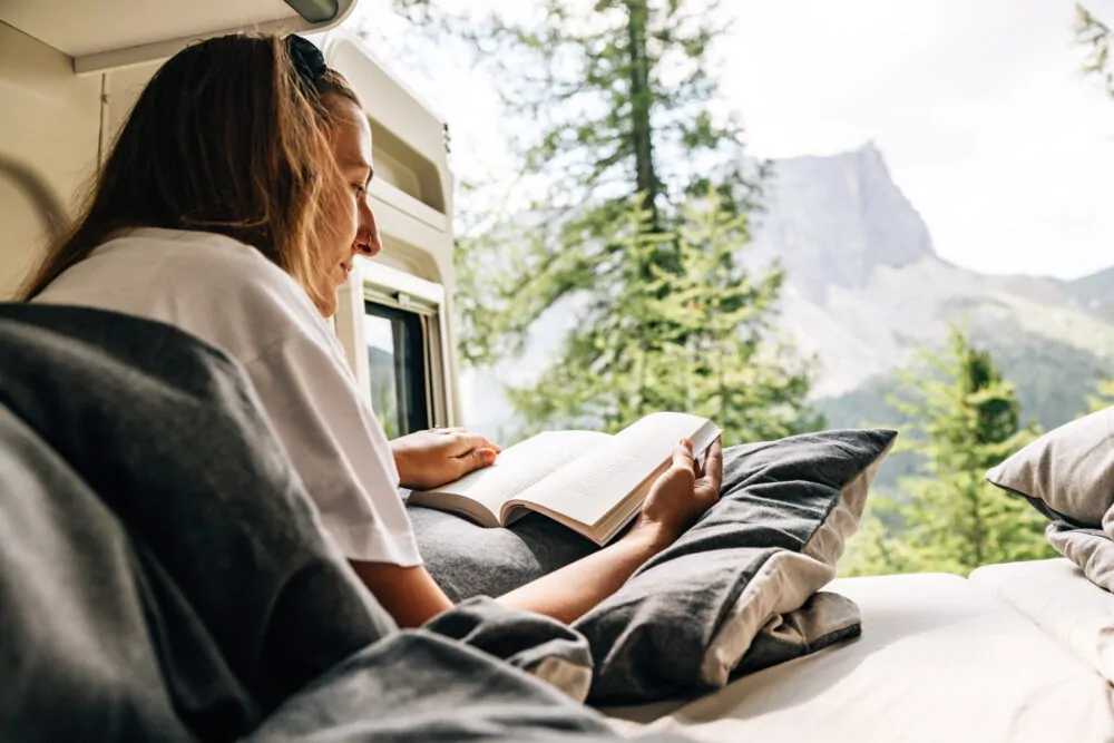 Eine Person liest ein Buch in einem Camper mit Blick auf einen bewaldeten Berg.