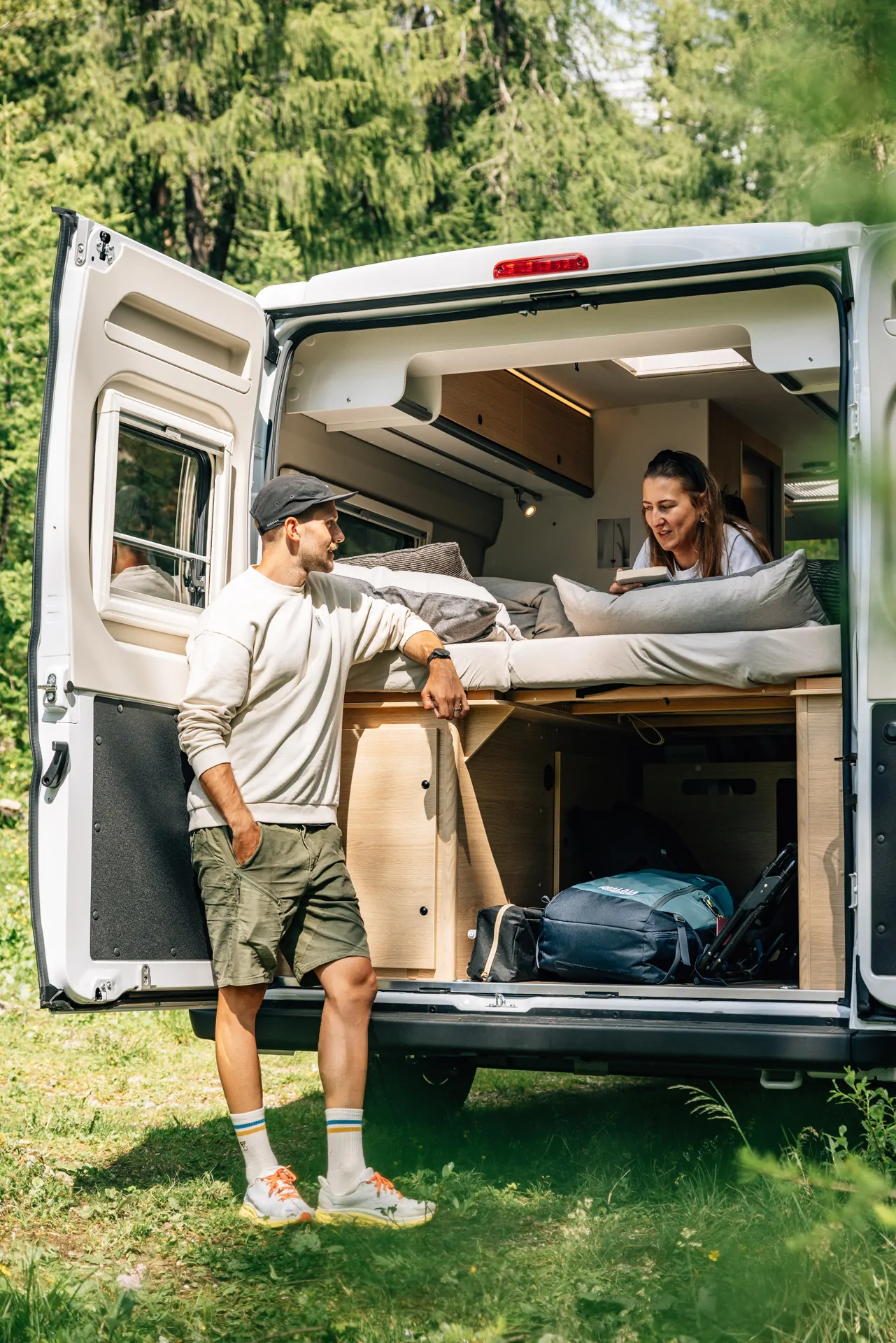 Ein Mann und eine Frau unterhalten sich am offenen Heck eines Sunlight Cliff X 2024 Campers im Grünen.