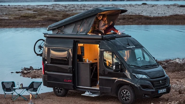Ein Sunlight Cliff RT Campervan an einem Seeufer, zwei Personen sitzen im Dachbereich.