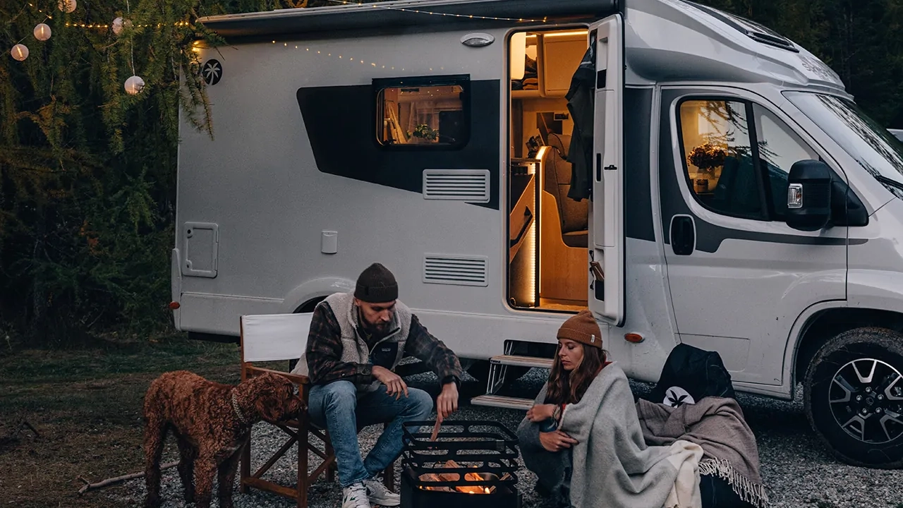 Zwei Personen sitzen mit einem Hund vor einem Wohnmobil am Lagerfeuer.