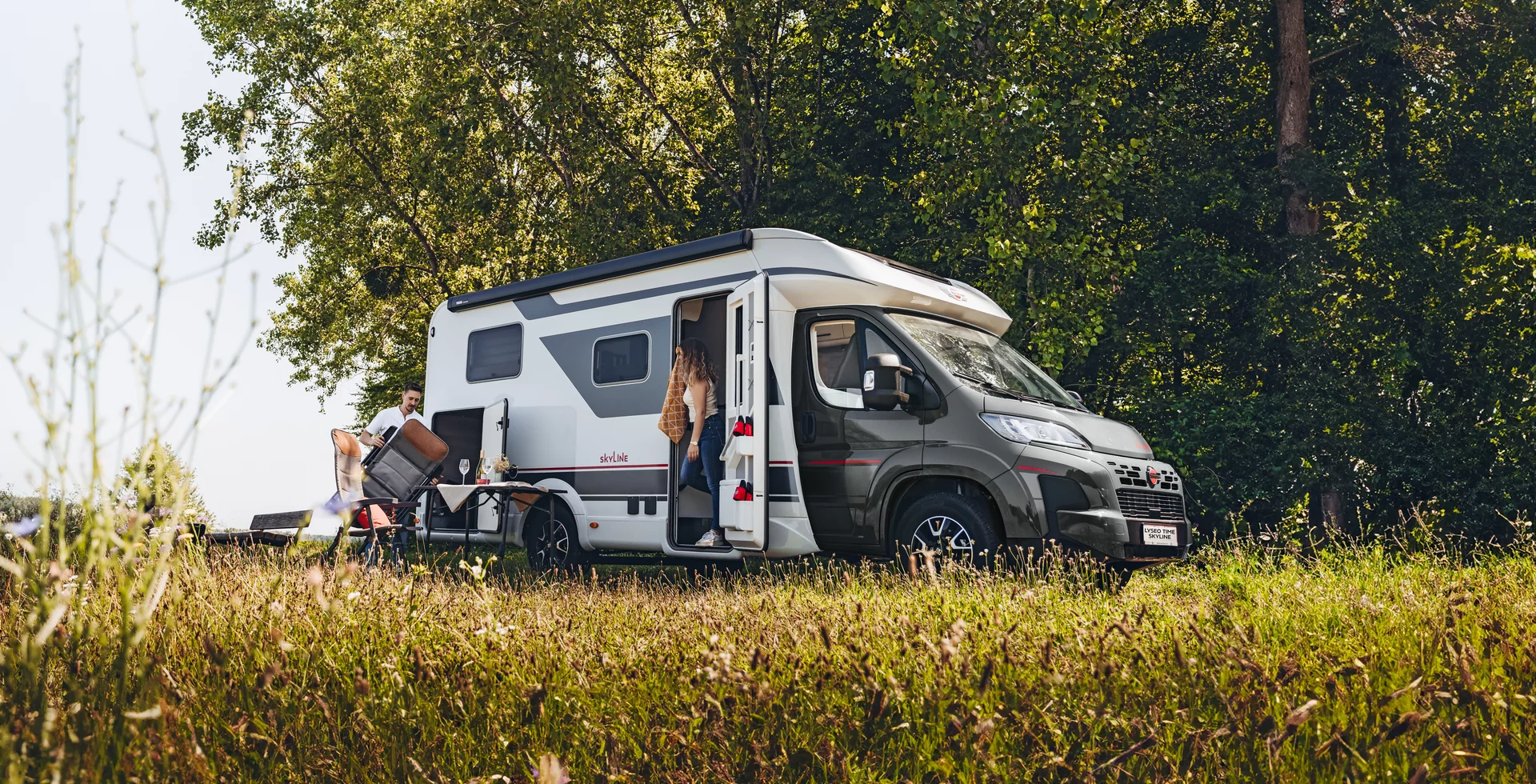 Ein Bürstner Lyseo Time Skyline Wohnmobil steht auf einer Wiese, während eine Person einsteigt und eine andere draußen sitzt.