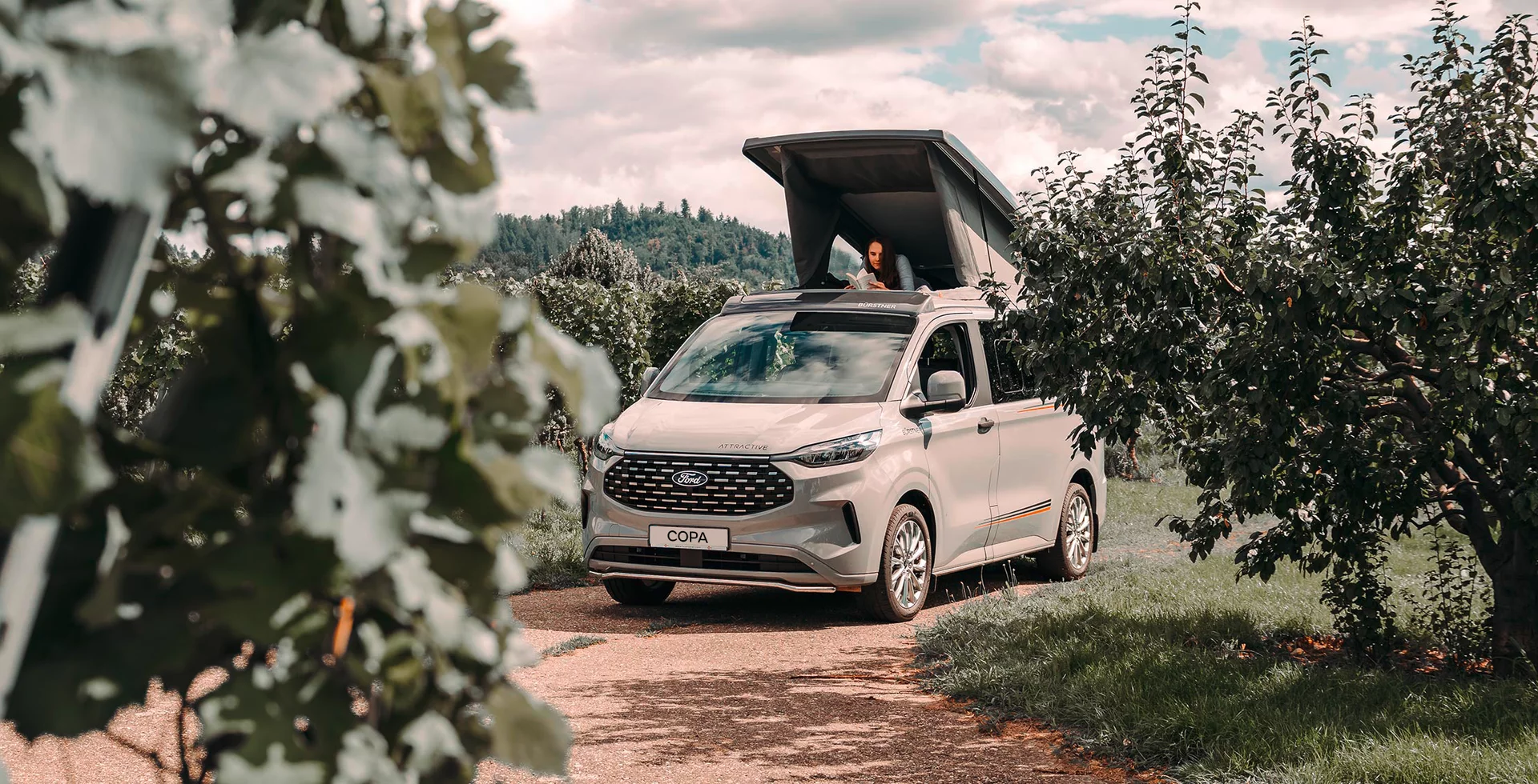 Ein Ford Bürstner Copa Camper mit geöffnetem Aufstelldach steht auf einem Weg zwischen Bäumen.