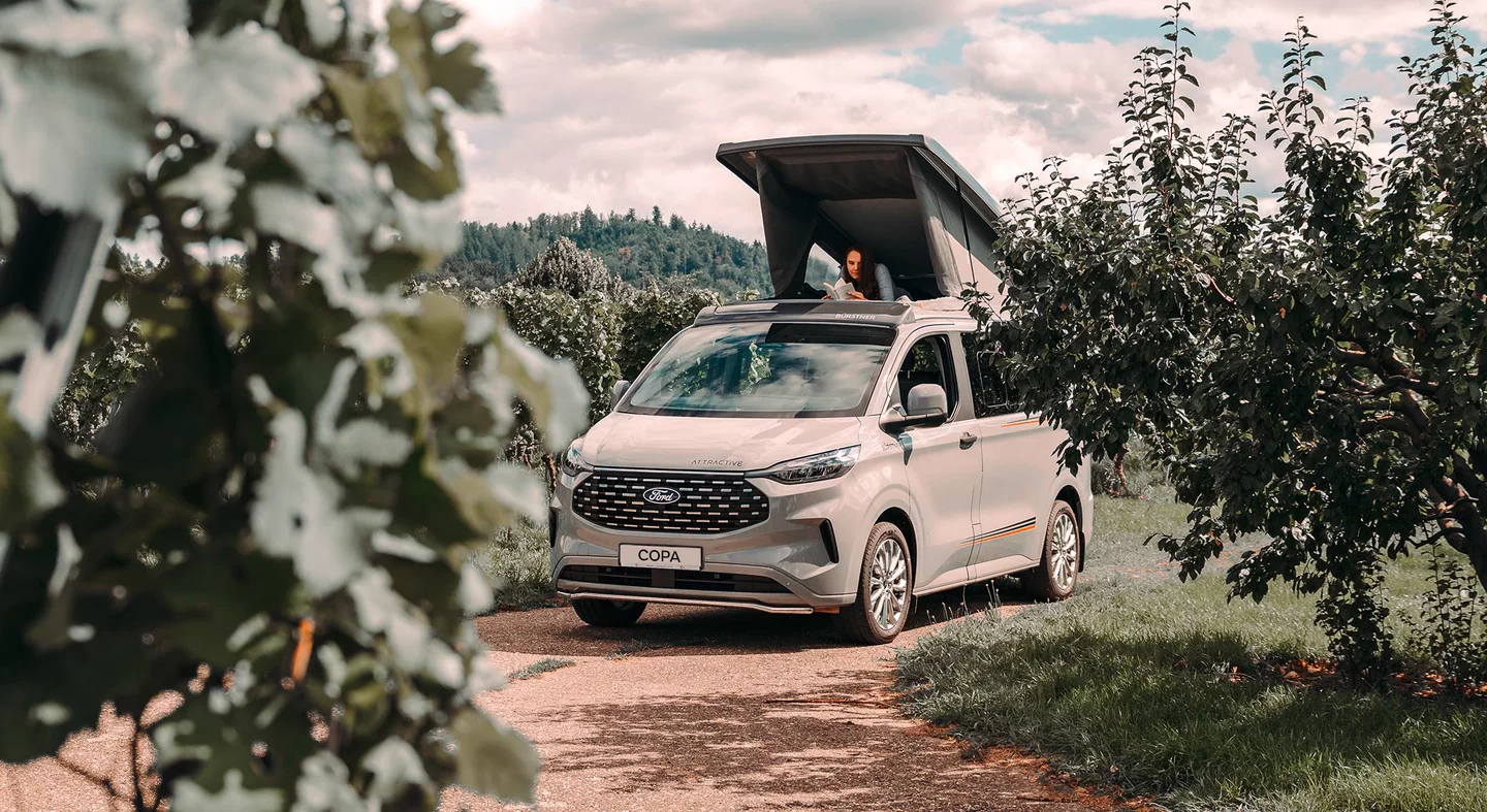 Ein Bürstner Copa Camper mit geöffnetem Aufstelldach steht auf einem Weg, umgeben von Bäumen.