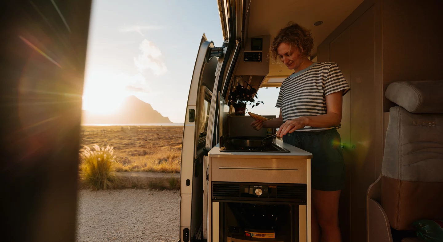 Eine Person kocht im Inneren eines Bürstner Delfin C621 Campers bei Sonnenuntergang.