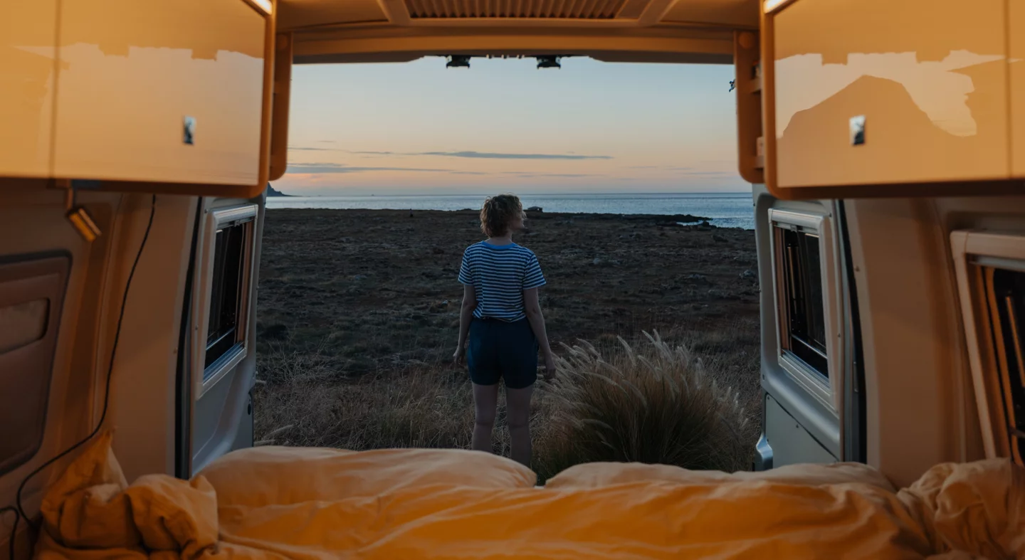 Eine Person steht in einem Bürstner-Camper mit Blick auf das Meer bei Sonnenuntergang.