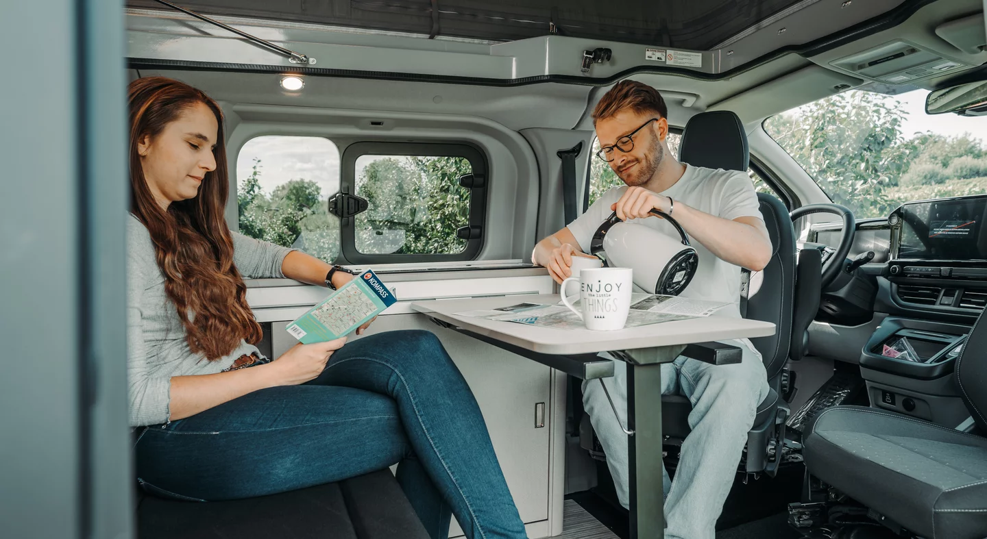 Zwei Personen sitzen in einem Bürstner Copa Wohnmobil, eine liest eine Karte, die andere gießt Tee ein.