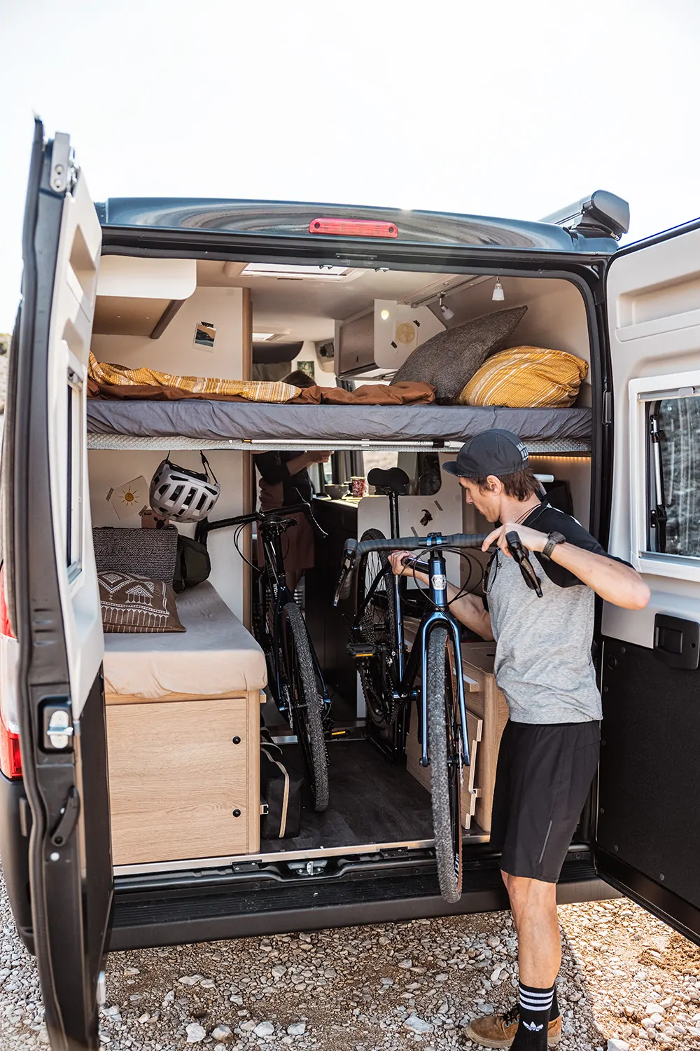 Ein Mann lädt Fahrräder in einen Camper mit geöffnetem Heck, in dem ein Bett und Stauraum sichtbar sind.