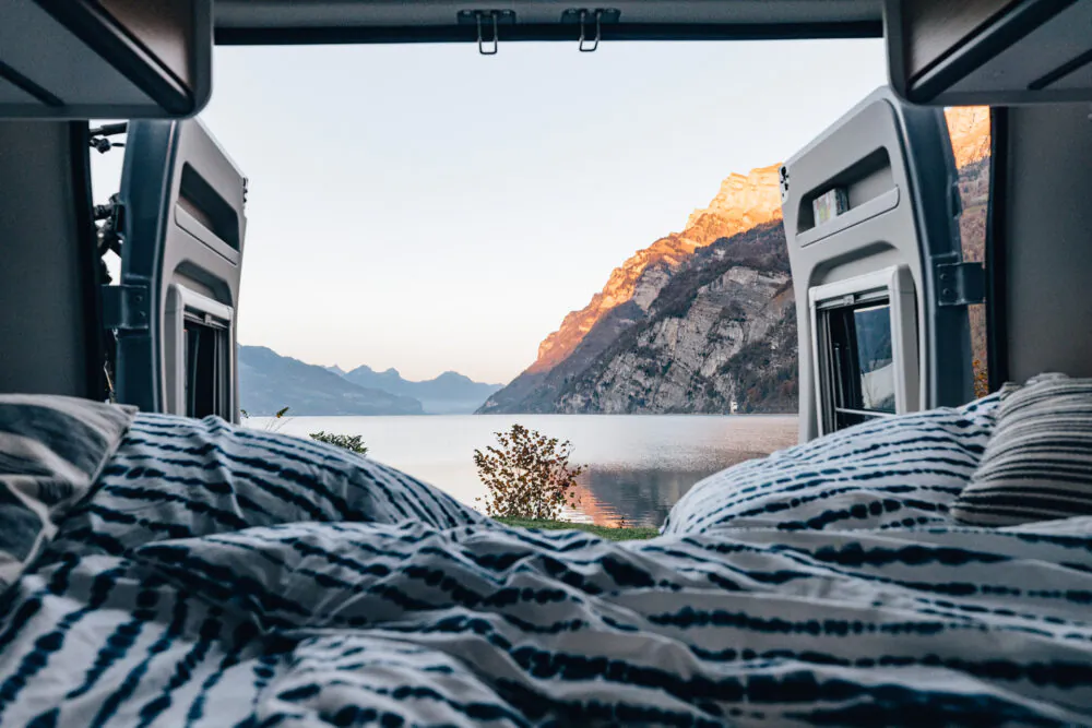 Blick aus einem Camper auf einen See und Berge im Sonnenuntergang.