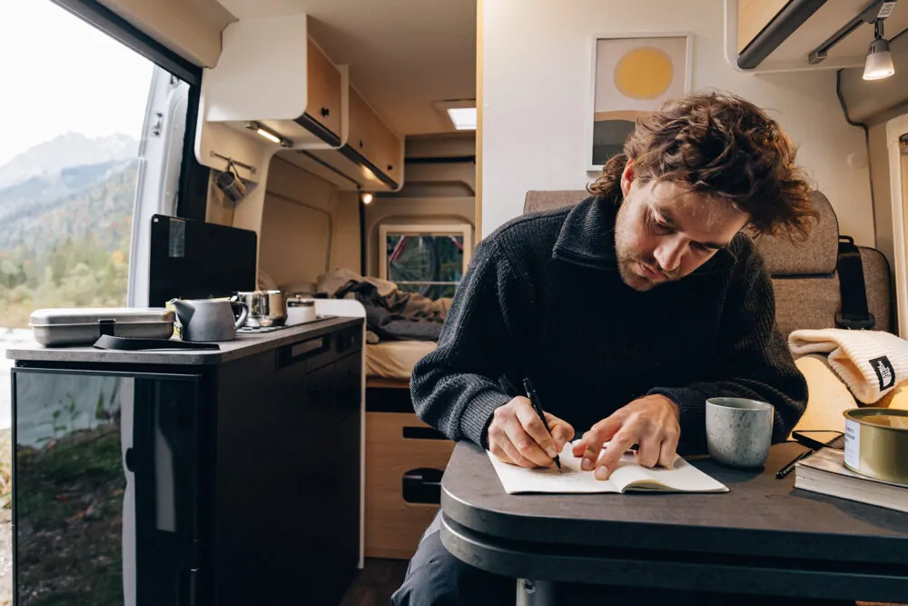 Ein Mann schreibt in einem Notizbuch am Tisch eines Peugeot Campervans, umgeben von einer gemütlichen Inneneinrichtung.