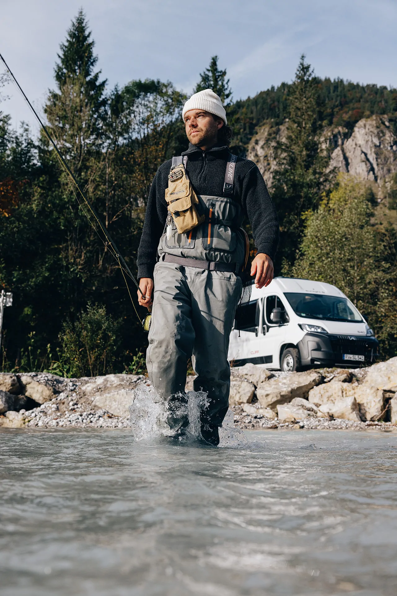 Ein Mann watet in Anglerkleidung durch einen Fluss, während im Hintergrund ein weißer Peugeot Sunlight Cliff Camper steht.