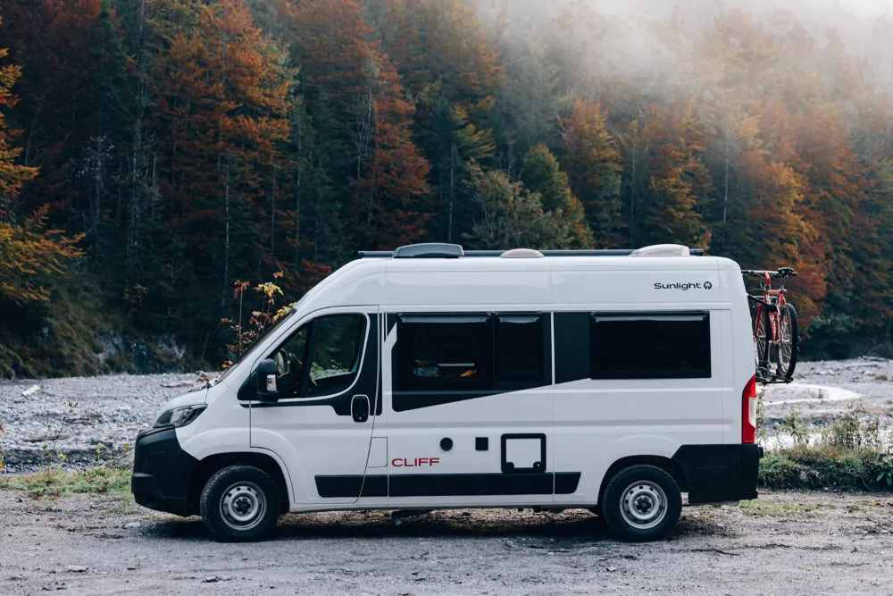 Ein weißer Sunlight Cliff Campervan mit einem Fahrradträger steht vor einem herbstlichen Wald.