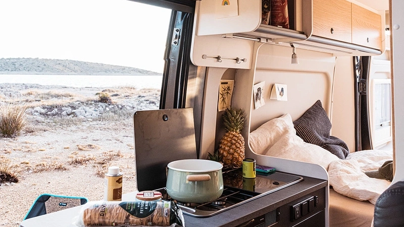 Innenansicht eines Campervans mit Küche und Schlafbereich, im Hintergrund eine Landschaft mit Wasser.
