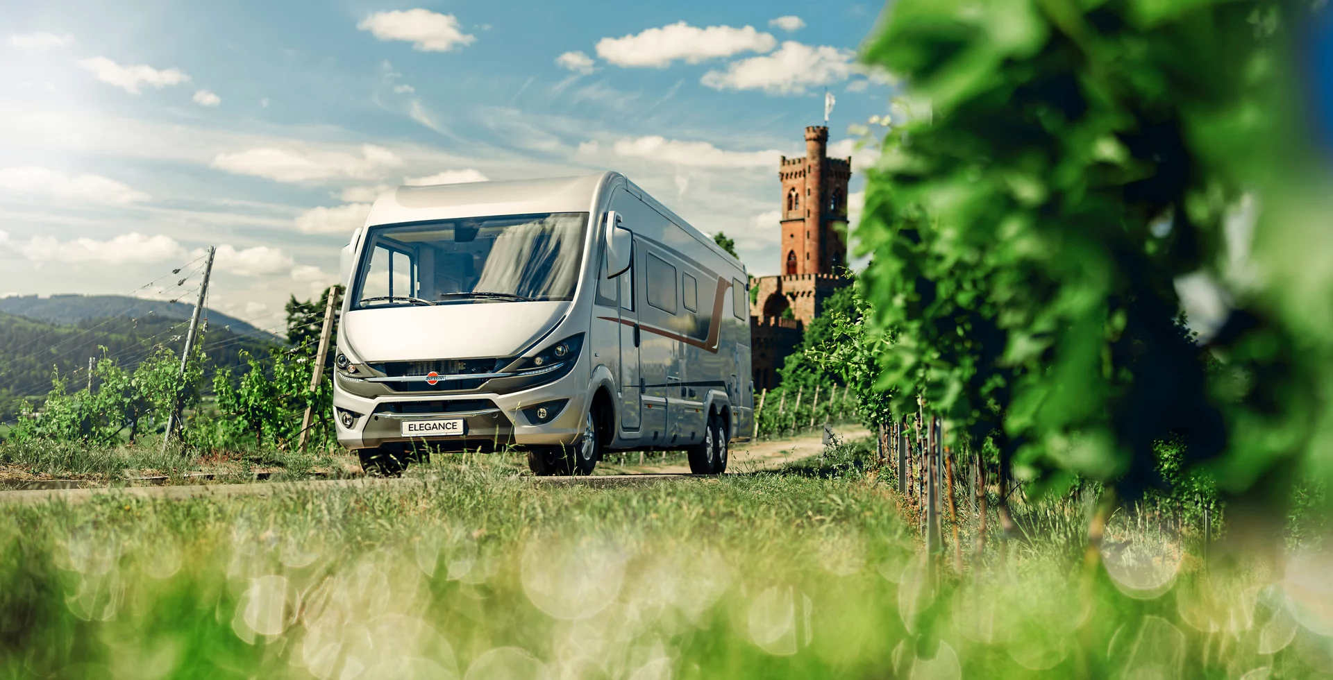 Ein Wohnmobil der Marke Bürstner Elegance fährt durch eine grüne Landschaft mit einem alten Turm im Hintergrund.