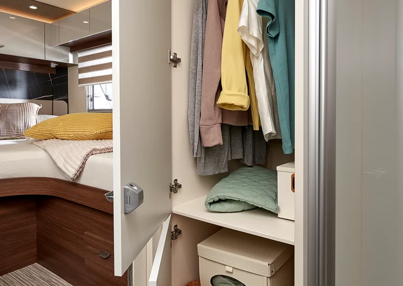 Offener Kleiderschrank in einem Wohnmobil mit hängender Kleidung und Bett im Hintergrund.