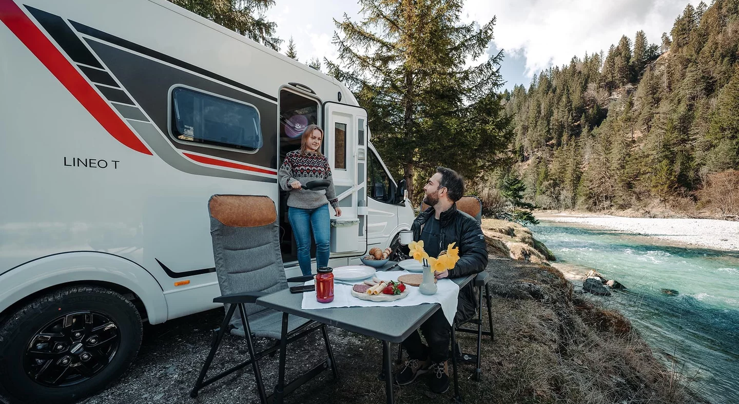 Ein Paar beim Frühstück vor einem Lineo T Wohnmobil in einer Wald- und Flusslandschaft.