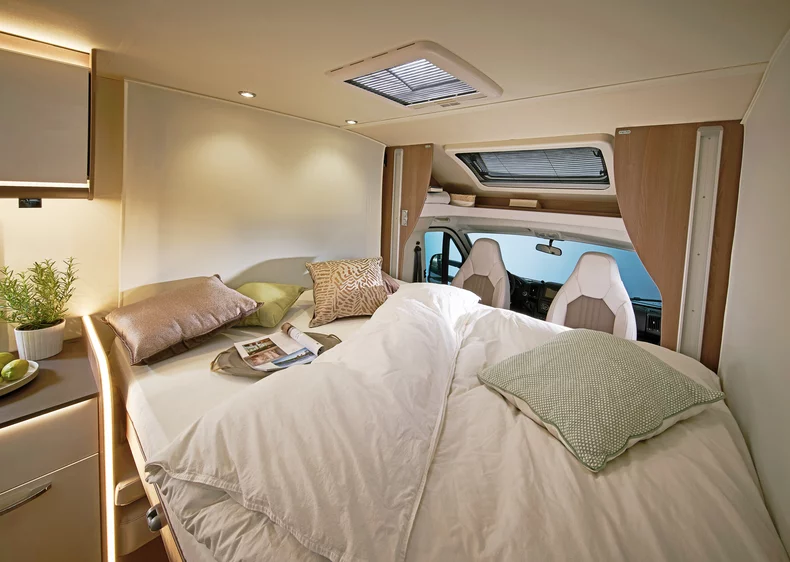 Innenansicht eines Wohnmobils mit einem gemachten Bett, dekorativen Kissen und einem Blick auf den Fahrerbereich.