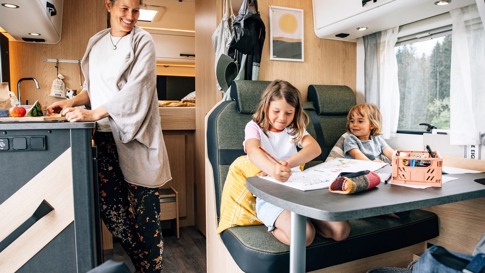 Eine Frau bereitet Essen in einem Wohnmobil vor, während zwei Kinder am Tisch malen.