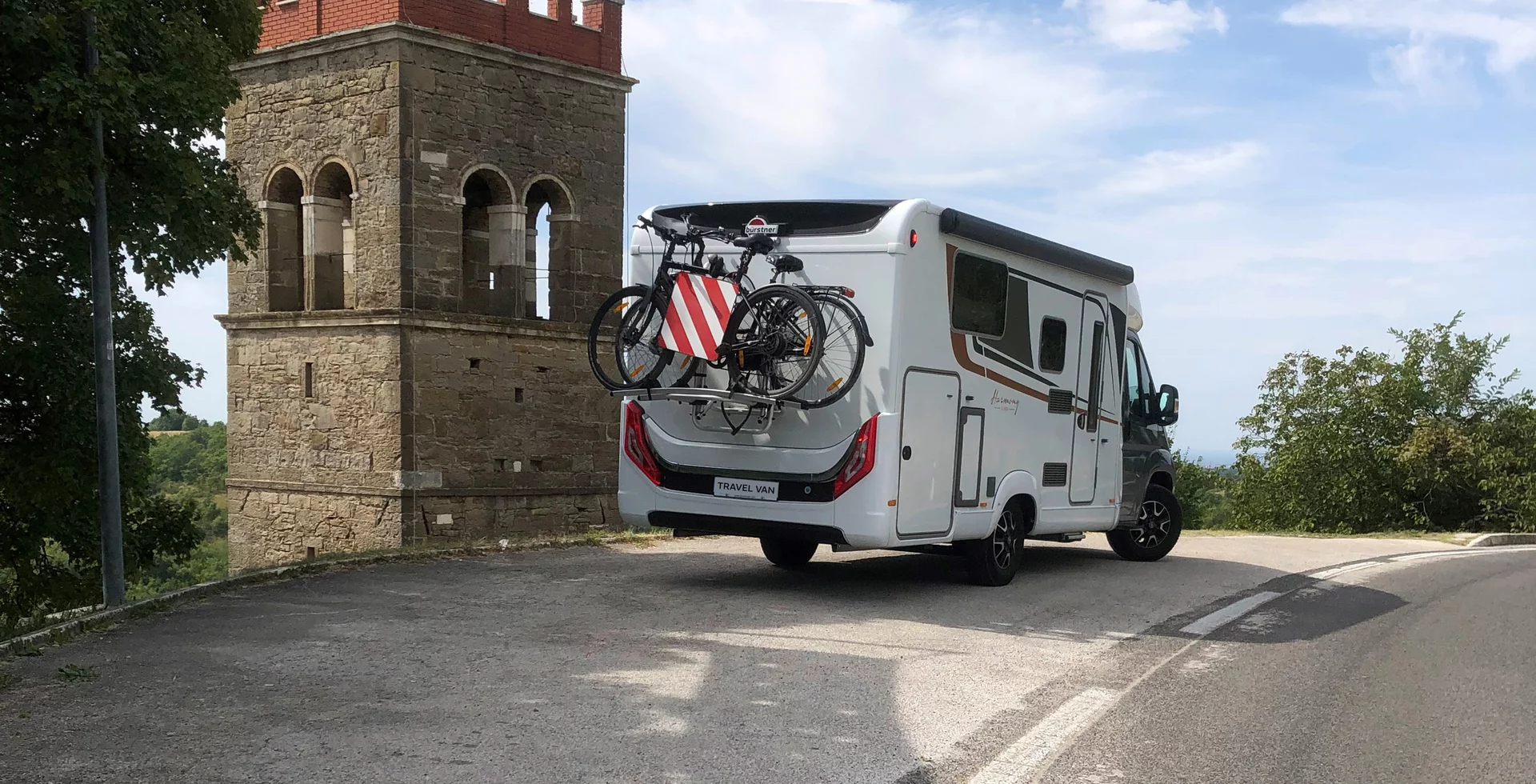 Ein Wohnmobil der Marke Bürstner mit zwei Fahrrädern am Heck steht neben einem alten Turm.