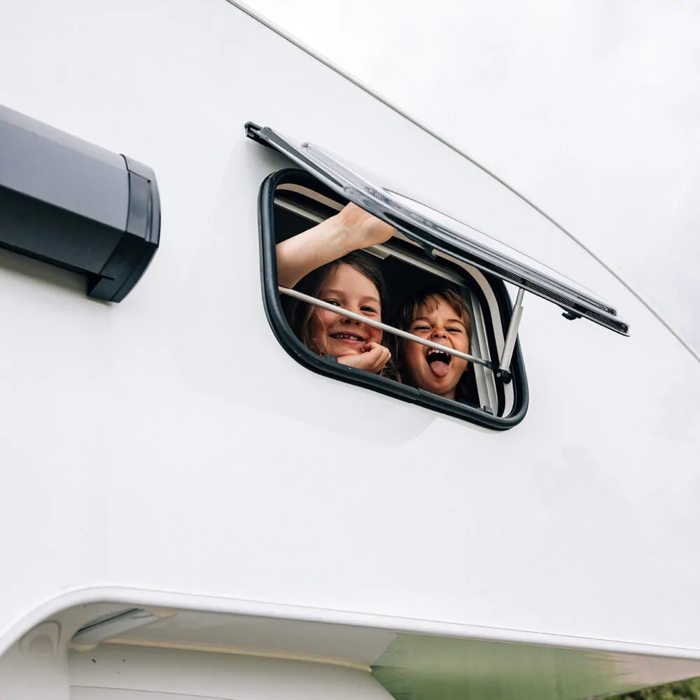 Zwei lachende Kinder schauen aus einem Fenster eines Wohnmobils.