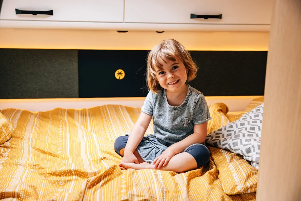Ein lächelndes Kind sitzt auf einem gelben Bett in einem Alkoven-Wohnmobil.