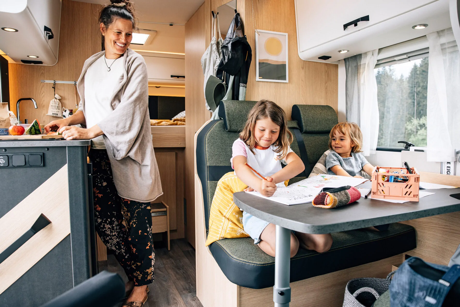 Eine Frau bereitet Essen zu, während zwei Kinder im Alkoven-Wohnmobil an einem Tisch malen.