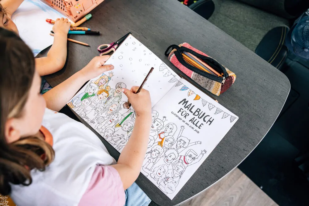 Ein Kind malt in einem Malbuch mit der Aufschrift "Malbuch für alle" an einem Tisch.