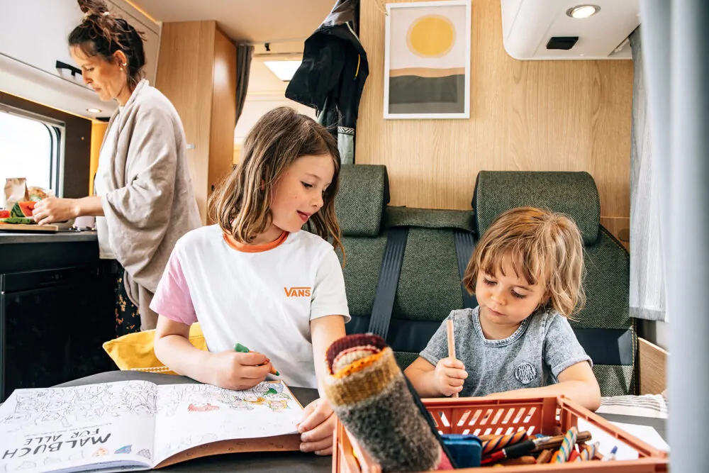 Eine Frau bereitet Essen zu, während zwei Kinder im Alkoven-Wohnmobil malen.