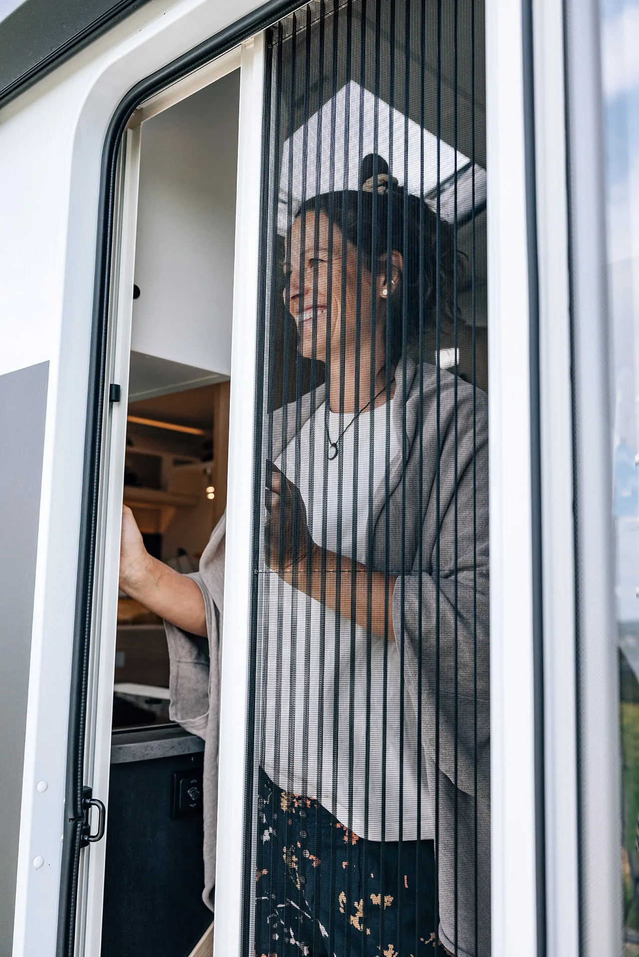 Eine Frau lächelt durch eine geöffnete Tür eines Campers mit Fliegengitter.