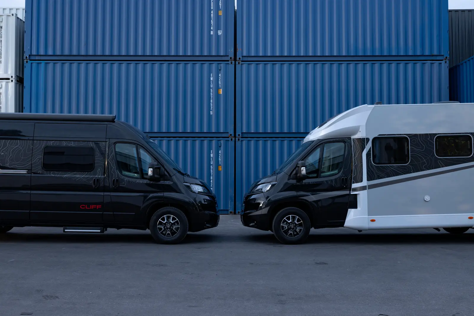 Zwei Campervans, darunter ein schwarzer mit der Aufschrift "CLIFF", stehen sich vor blauen Containern gegenüber.