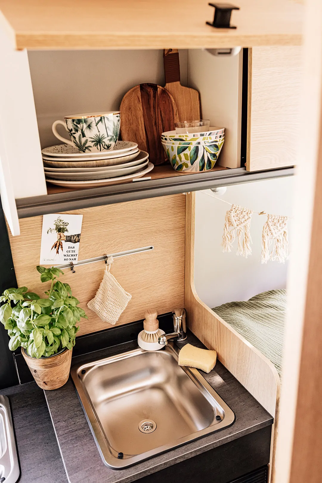 Küchenbereich in einem Camper mit Spüle, Basilikumtopf und Geschirr im Schrank.