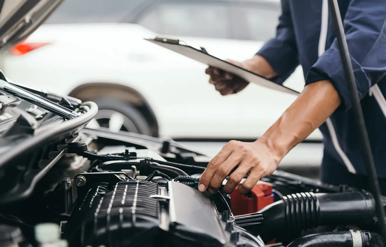 Eine Person inspiziert den Motorraum eines Autos mit einem Klemmbrett in der Hand.