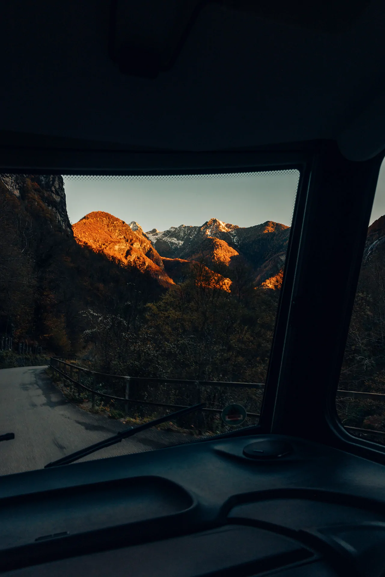 Blick aus einem Fahrzeug auf eine kurvige Straße und sonnenbeschienene Berge im Hintergrund.