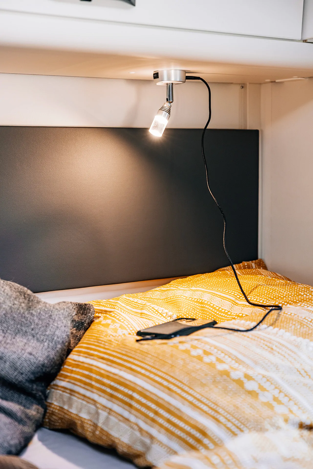 Eine Leselampe über einem Bett mit gelbem Kissen und einem Smartphone.