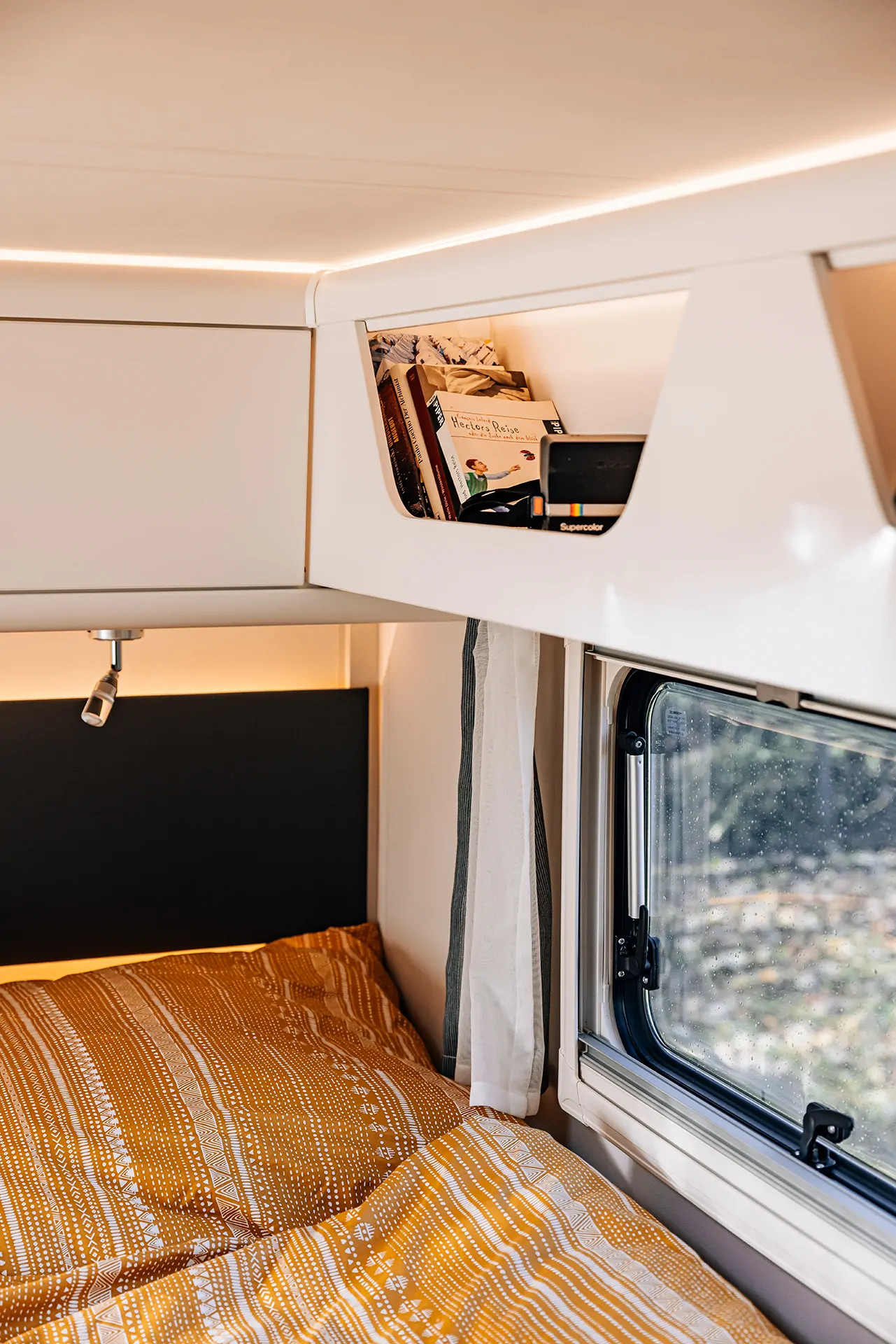 Ein gemütliches Bett mit orange gemustertem Bettbezug neben einem Fenster und einem Regal mit Büchern in einem Wohnmobil.