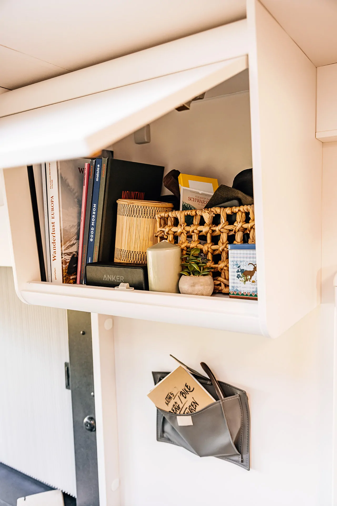 Ein offenes Regal in einem Camper zeigt Bücher, eine Kerze, eine Pflanze und eine kleine Tasche mit Karten.